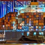 A brightly lit cargo ship at Hamburg harbor with stacked containers and a tugboat.