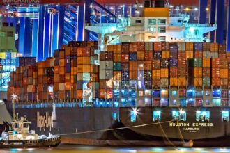 A brightly lit cargo ship at Hamburg harbor with stacked containers and a tugboat.