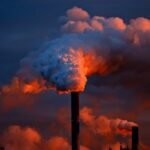Smoke billows from industrial chimneys at sunset, highlighting pollution against a vibrant sky.