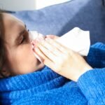 A woman in a blue sweater uses a tissue, lying indoors, suggesting illness or flu symptoms.
