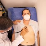 Doctor in protective mask examines patient with stethoscope in medical clinic.