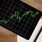 Flatlay of an iPad displaying stock market graph on a wooden desk with a pencil and paper.