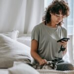 Concentrated young ethnic woman with curly hair in casual clothes messaging on mobile phone and stroking cat while relaxing on comfortable bed during weekend
