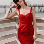 Elegant woman in a red polka dot dress enjoys a sunny day at the beach pier, showcasing vibrant summer fashion.