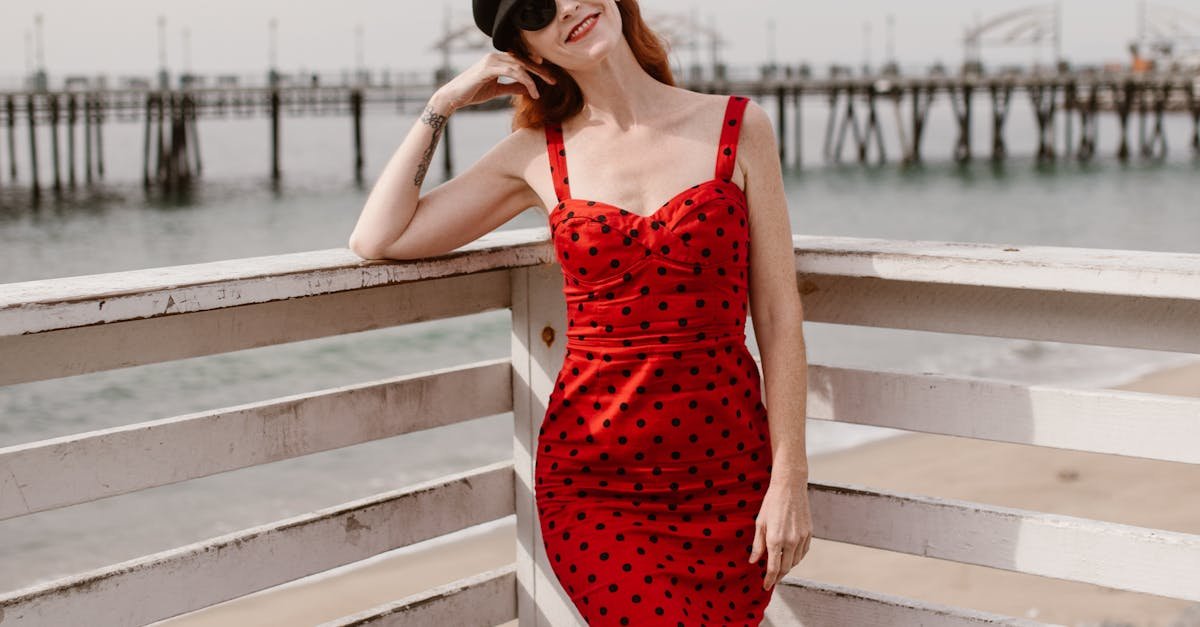 Elegant woman in a red polka dot dress enjoys a sunny day at the beach pier, showcasing vibrant summer fashion.