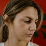 Close-up portrait of a woman with a bruised eye looking down, conveying emotion against a red backdrop.