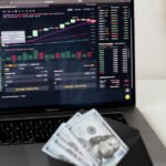 A person examines stock market data on a laptop while holding dollar bills, showcasing finance and trading concepts.