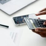 A businessman using a laptop and smartphone to analyze stock market trends indoors.