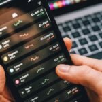 Hands holding smartphone with stock app, laptop displaying charts. Perfect for financial and technology themes.