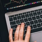 Hands typing on a laptop with cryptocurrency charts and smartphone display in view, focusing on digital finance.