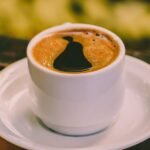Steaming cup of black coffee placed on a saucer with blurred background.