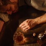 A man sleeps on a table with a whiskey glass and bottle nearby, suggesting exhaustion.