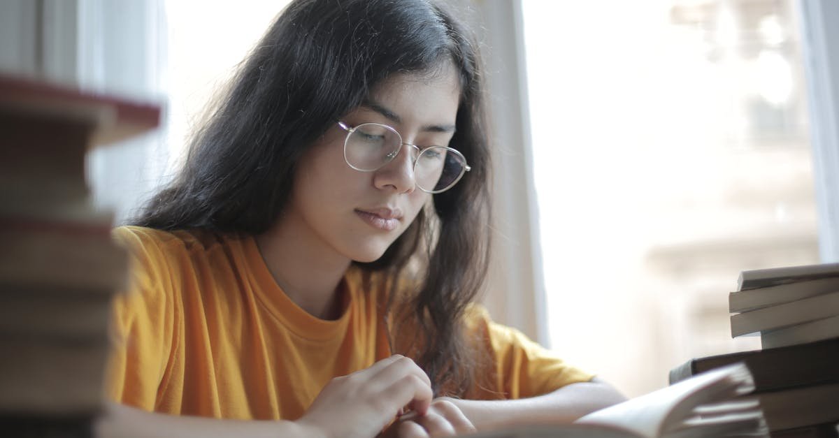 Low angle of diligent ethnic female student wearing casual t shirt and eyeglasses sitting at table with stacks of books and preparing for exam