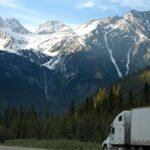 A semi-truck travels along a highway with snow-capped mountains in the background.