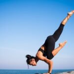 Woman performing a yoga pose on a rocky beach with ocean view and clear blue sky.