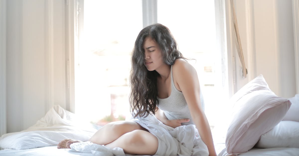 Woman sitting on bed holding her stomach, showing signs of discomfort or pain indoors.