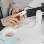 Adult using a pulse oximeter at a home desk, surrounded by tech and healthcare items.