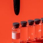 Black gloved hand pointing at a row of transparent vaccine vials on red background.